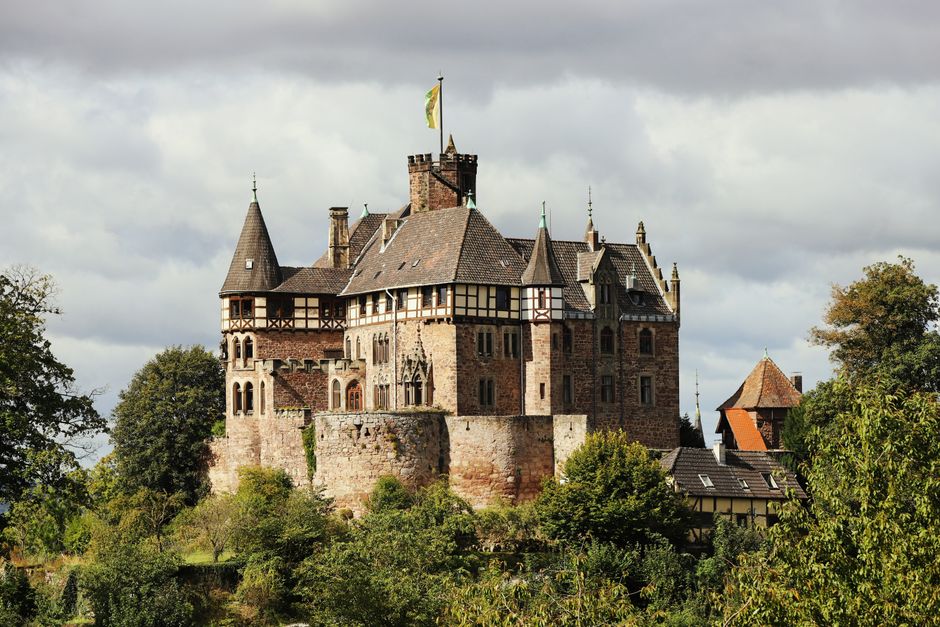 Photograph of a historic medieval castle situated on a hill surrounded by lush green trees. The castle features multiple towers with conical roofs, battlements, and a variety of stone and timber construction elements. The weather appears overcast with grey clouds overhead, providing diffused lighting that highlights the intricate stonework and architectural details of the structure. At the base of the hill, there are residential buildings with slate and tile roofs. The overall scene suggests a peaceful setting, possibly in a rural area, with the castle serving as a prominent landmark. Although the image does not depict any moving activity or furniture, it visually supports themes of historical architecture relevant to house and property relocation services often associated with historic properties, such as those handled by Man and Van Earls Court during house removals or furniture transport for listings and estate management.