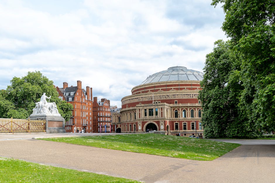 A wide-angle view of a historic theatre within an outdoor park setting, featuring a large, rounded brick and stone building with arched windows and a domed roof, surrounded by lush green trees and well-maintained grass. To the left, there is a white stone statue depicting multiple figures, mounted on a pedestal, positioned atop a paved area adjacent to the grass. In the foreground, a gravel pathway runs through the park, leading towards the theatre entrance, with a loading area visible near the building. The scene is illuminated by diffused daylight under a partly cloudy sky, capturing the process of an outdoor home relocation or furniture transport involving loading or unloading at a nearby vehicle or van, with some moving equipment such as trolleys or straps potentially present but not clearly visible. Man and Van Earls Court occasionally operate in areas like this, handling packing and moving logistics for local house removals.
