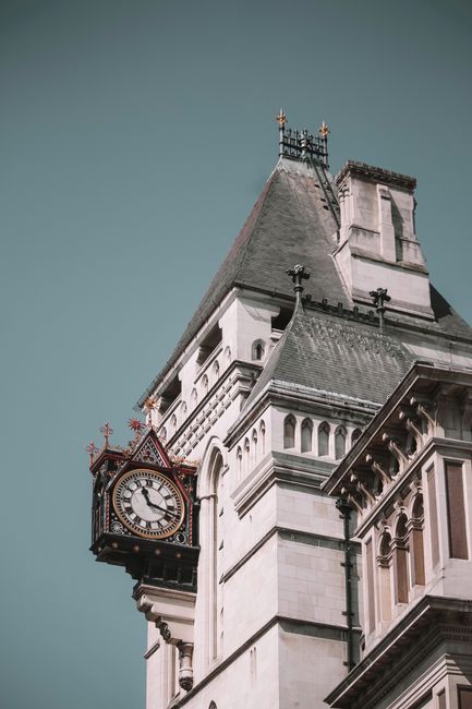 This image shows a close-up view of a historic clock tower on a large stone building, likely part of a government or civic structure. The clock face is ornate, with Roman numerals and decorative gold accents, and is mounted on a protruding section of the tower. The structure features detailed architectural elements, including arched windows, decorative cornices, and stone carvings, with a steep, slate-tiled roof topped by a small spire. The building appears to be in a city setting, with the sky in the background. This scene may be relevant to home relocation or moving services, such as those provided by Man and Van Earls Court, highlighting the importance of careful planning and logistics in furniture transport and packing during house removals or office moves.