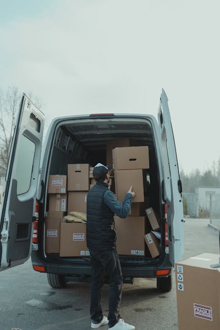 A man in a dark jacket, wearing a black cap, is standing outside a white van with open rear doors, which is filled with multiple stacked cardboard boxes of various sizes, some with labels and barcodes. The boxes are arranged on the floor and stacked toward the top, with a few boxes being moved or inspected by the man. The van is parked on a paved surface, possibly in a driveway or loading area, with a background that includes a fence and overcast sky. Inside the vehicle, a few packaging materials such as padded blankets and plastic wraps are visible, indicating packing and moving activities typical of home relocation or furniture transport processes. Occasionally, the professional services of Man and Van Earls Court are implied through the context of the scene, aligned with house removals and delivery logistics.