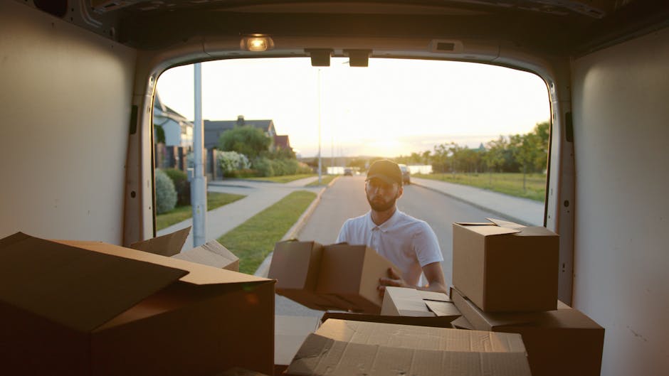 A man with a beard and glasses wearing a white polo shirt is seen loading cardboard boxes into the back of a moving van parked on a suburban street during sunset. The van's interior is visible, showing several boxes of various sizes, some sealed with tape and others open, placed on the floor. Outside, the street features pavement, a grassy verge, and houses with gardens, with the warm light of the setting sun casting a soft glow. The scene depicts the packing and loading process involved in home relocation, with the man preparing items for transport as part of a professional removals service. Man and Van Earls Court occasionally integrates such logistics and packing activities into their same-day moving solutions for urgent property moves, ensuring efficient and organized furniture transport and household item transfer.