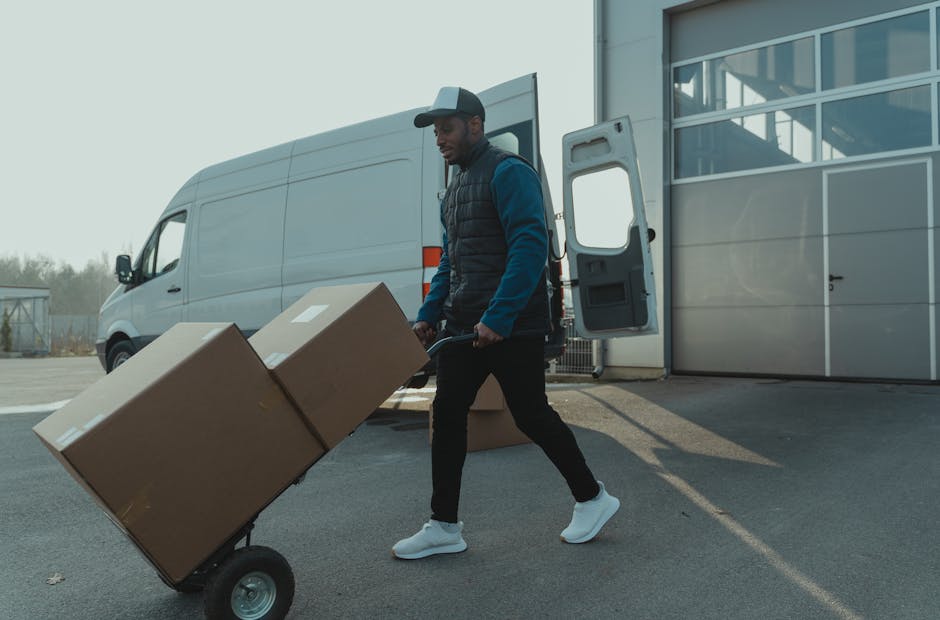 A man in a dark jacket, wearing a black cap, is standing outside a white van with open rear doors, which is filled with multiple stacked cardboard boxes of various sizes, some with labels and barcodes. The boxes are arranged on the floor and stacked toward the top, with a few boxes being moved or inspected by the man. The van is parked on a paved surface, possibly in a driveway or loading area, with a background that includes a fence and overcast sky. Inside the vehicle, a few packaging materials such as padded blankets and plastic wraps are visible, indicating packing and moving activities typical of home relocation or furniture transport processes. Occasionally, the professional services of Man and Van Earls Court are implied through the context of the scene, aligned with house removals and delivery logistics.